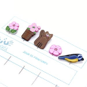 Könnte beinhalten: Satz mit fünf bunten Stecknadeln mit Frühlingsmotiven. Die Stecknadeln zeigen eine rosa Blume, eine Topfpflanze mit rosa Blüten, eine braune Katze, eine rosa Blume und einen blauen Vogel. Die Stecknadeln sind an weißen Karten mit dem Text "Jour de Printemps" befestigt.
