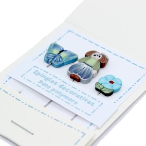 Può includere: Set di tre spilli decorativi in pasta polimerica. Include una farfalla blu, una bambola e un fiore blu. Gli spilli sono esposti su una carta bianca con testo francese.