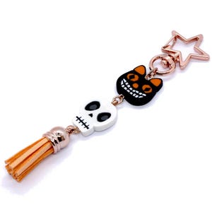Puede incluir: Un llavero temático de Halloween con una calavera blanca, un gato negro con ojos naranjas y un cierre en forma de estrella. El llavero tiene una cadena dorada y una borla de color marrón claro.