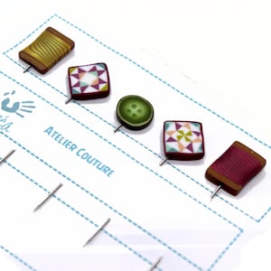 Könnte beinhalten: Ein Set mit fünf bunten Nähten, darunter eine Garnrolle, ein Knopf und zwei Patchwork-Quadrate. Die Pins werden auf einer weißen Karte mit dem Text "Atelier Couture" angezeigt.