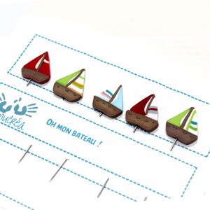 Könnte beinhalten: Fünf bunte, segelschiffartige Stecknadeln mit braunen Sockeln. Die Stecknadeln sind in einer Reihe auf einer weißen Karte mit blauen gestrichelten Linien und dem Text "Oh mon bateau!" angeordnet.