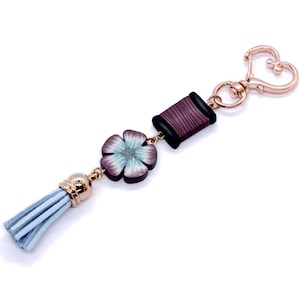 Puede incluir: Un llavero decorativo con una borla azul claro, una flor con pétalos azules y morados y una bobina de hilo. El llavero tiene un cierre en forma de corazón y detalles dorados. La flor tiene un pequeño centro plateado.