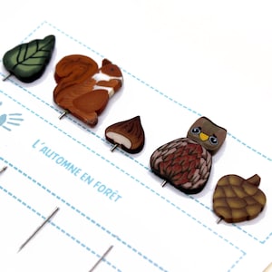 Könnte beinhalten: Ein Set aus fünf Holzstiften mit Herbstmotiven. Die Stifte zeigen ein Eichhörnchen, eine Kastanie, eine Eule, eine Tannenzapfen und ein Blatt. Die Stifte befinden sich auf einer weißen Karte mit dem Text "L'automne en forêt".