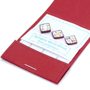 Könnte beinhalten: Ein rotes Stoffetui enthält drei dekorative Polymer Clay Pins. Jeder quadratische Pin hat ein geometrisches Design in Pastellfarben. Die Pins befinden sich auf einer weißen Karte mit französischem Text.