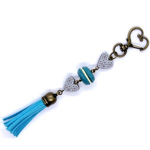 Puede incluir: Un llavero decorativo con una borla azul y un cierre en forma de corazón de color bronce. El llavero presenta un dije de macaron azul y dos dijes de corazón grises con un patrón geométrico. Los detalles metálicos son de color bronce.