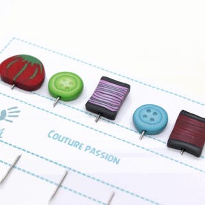 Può includere: Spilli da cucito fatti a mano con decorazioni. Le decorazioni includono un pomodoro rosso, un bottone verde, una bobina di filo viola, un bottone blu e una bobina di filo marrone. Su una carta bianca con il testo "Couture Passion".