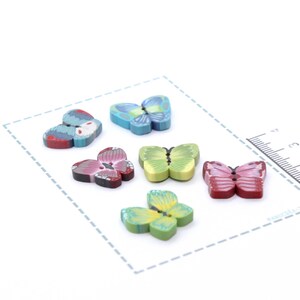 Peut inclure: Six boutons décoratifs en forme de papillon, aux couleurs vives : bleu, vert, rose et rouge. Ils sont disposés sur une surface blanche avec un bord pointillé et une règle graduée pour la mesure.