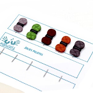 Könnte beinhalten: Fünf bunte Wollknäuel mit schwarzen Mittelpunkten, jeweils an einer Stecknadel befestigt, werden auf einer weißen Karte mit dem Text "Jolies Pelotes" und einem handgezeichneten Logo präsentiert.