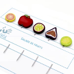 Könnte beinhalten: Förmige Anstecknadeln mit einer roten Apfel, einer Passionsfrucht, einer Feige und einer Zitronenscheibe. Die Nadeln sind auf einer weißen Karte mit dem Text "Salade de Fruits" angeordnet.