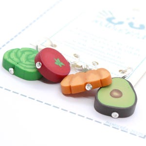 Può includere: Un set di quattro segnapunti a forma di cibo composto da una foglia di lattuga verde, una fetta di pomodoro rosso, una zucca arancione e un avocado tagliato a metà con nocciolo marrone, fissati a piccoli anelli metallici color argento.