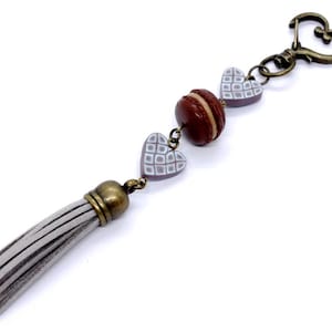 Puede incluir: Un llavero decorativo con un dije de macaron marrón, dos dijes en forma de corazón y una borla gris. El llavero tiene un cierre y herrajes de conexión de color bronce. Los dijes de corazón tienen un patrón de cuadrícula blanca.