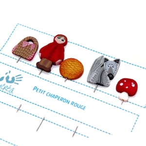 Könnte beinhalten: Ein Set mit fünf bunten Pins, die Figuren aus der Geschichte von Rotkäppchen zeigen. Die Pins beinhalten Rotkäppchen, einen Wolf, einen Korb, einen Pilz und ein Brot. Die Pins sind aus Polymer Clay gefertigt und an Metallnadeln befestigt.