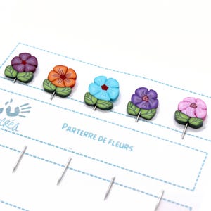 May include: Five colourful flower-shaped pins with green leaves. The flowers are in shades of purple, orange, blue, and pink. The pins are arranged on a white card with the text "PARTERRE DE FLEURS".