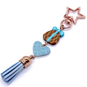Puede incluir: Un llavero decorativo con un cierre en forma de estrella y una borla azul claro. El llavero presenta un dije en forma de regalo marrón y azul, un dije de corazón azul y un anillo de metal dorado.