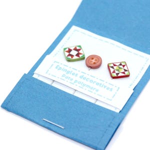 Könnte beinhalten: Ein blaues Filzetui enthält dekorative Anstecknadeln und einen Knopf. Zwei quadratische Nadeln zeigen ein patchworkartiges Design in Rot, Grün und Weiß. Ein runder, hellbrauner Knopf ist ebenfalls enthalten. Der Text "Epingles décoratives Pate polymère" ist auf eine weiße Karte gedruckt.