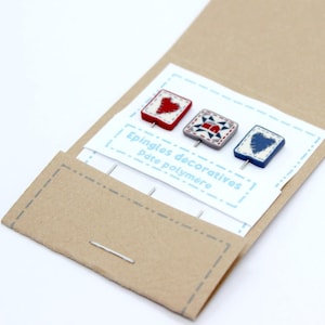 Könnte beinhalten: Drei dekorative Polymer Clay Pins mit quadratischen Designs, darunter ein rotes Herz, ein geometrisches Muster und ein blaues Herz. Die Pins werden auf einer weißen Karte mit französischem Text in einer braunen Papierverpackung präsentiert.