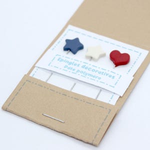 Peut inclure: Un ensemble d'épingles décoratives dans un emballage en papier kraft. Les épingles présentent une étoile bleue, une étoile blanche et un cœur rouge. Le texte "Epingles décoratives Pâte polymère" est imprimé sur la carte.