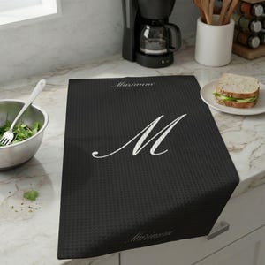Op de afbeelding: Een zwarte keukenhanddoek met wafelstructuur en de witte letter "M". De naam "Marianne" staat in sierlijke letters boven en onder de letter. De handdoek ligt op een aanrecht.