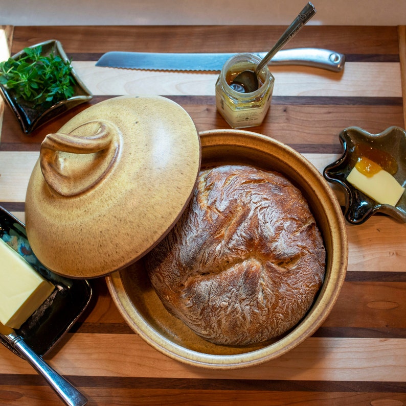 May include: A round, brown ceramic bread cloche with a lid and a loaf of freshly baked bread inside. The cloche is sitting on a wooden cutting board with a small dish of butter and a jar of honey.