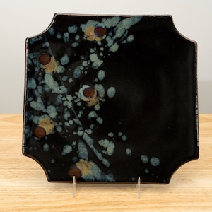 May include: A black square ceramic plate with a scalloped edge. The plate has a speckled glaze with blue, brown, and yellow accents.