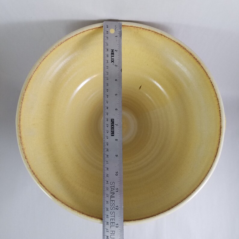May include: A large, yellow ceramic bowl with a slightly rippled surface and a brown rim. The bowl is empty and appears to be handmade.
