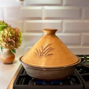 May include: A brown ceramic tagine with a decorative design of grass on the lid, sitting on a black gas stovetop with a blue flame.