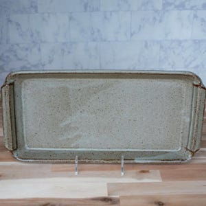 May include: A rectangular ceramic serving tray with a speckled, light gray finish. The tray has slightly raised edges and small handles on each end. The tray is displayed on a wooden surface.