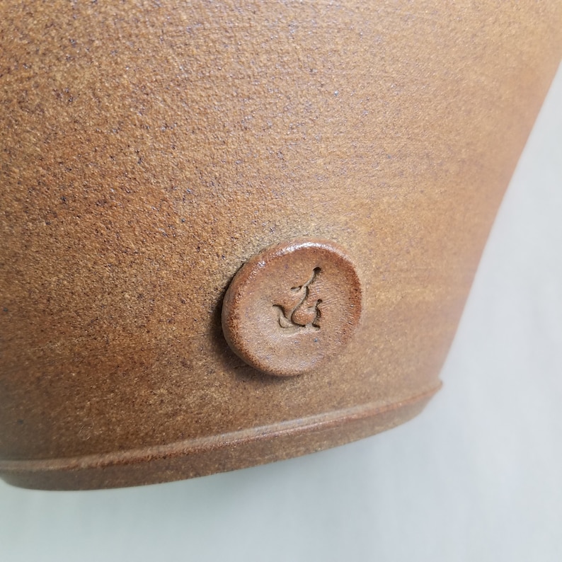 May include: Close-up of a brown ceramic pot with a round, embossed seal on the side. The seal features a stylised bird design.
