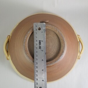 May include: A brown ceramic bowl with two handles. The bowl has a white rim and a brown interior. The bottom of the bowl is stamped with the text "OTTERY" and "L 4 MM". A ruler is placed next to the bowl for scale.
