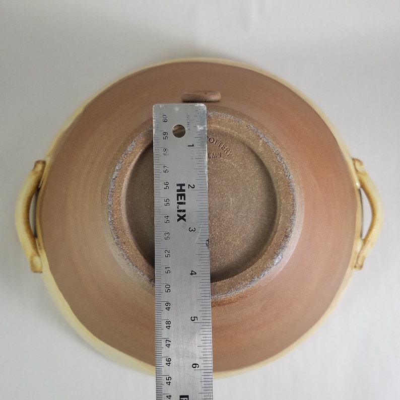 May include: A brown ceramic bowl with two handles. The bowl has a white rim and a brown interior. The bottom of the bowl is stamped with the text "OTTERY" and "L 4 MM". A ruler is placed next to the bowl for scale.