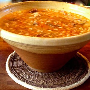 May include: A large, brown earthenware bowl filled with a creamy white bean soup with meat and vegetables. The bowl is sitting on a woven brown and white placemat.