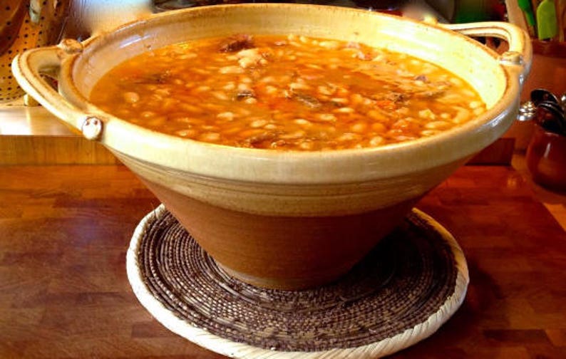 May include: A large, brown earthenware bowl filled with a creamy white bean soup with meat and vegetables. The bowl is sitting on a woven brown and white placemat.