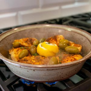 May include: A ceramic bowl filled with cooked chicken, green olives, and a lemon slice. The dish is seasoned with herbs and spices. The bowl is on a gas stove with a blue flame.