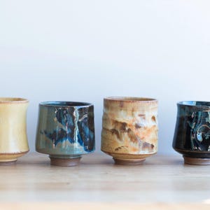 May include: Four handcrafted ceramic cups in a row. The cups have unique shapes and colors, including yellow, teal, brown, and dark blue. Each cup has a textured surface and sits on a wooden surface.