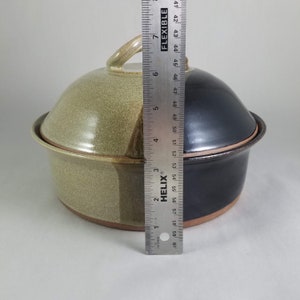 Pu&ograve; includere: Una pirofila in ceramica bicolore con coperchio. Il lato sinistro &egrave; beige e il destro &egrave; nero. Il coperchio ha una maniglia. Un righello mostra che il piatto &egrave; alto circa 15 cm.