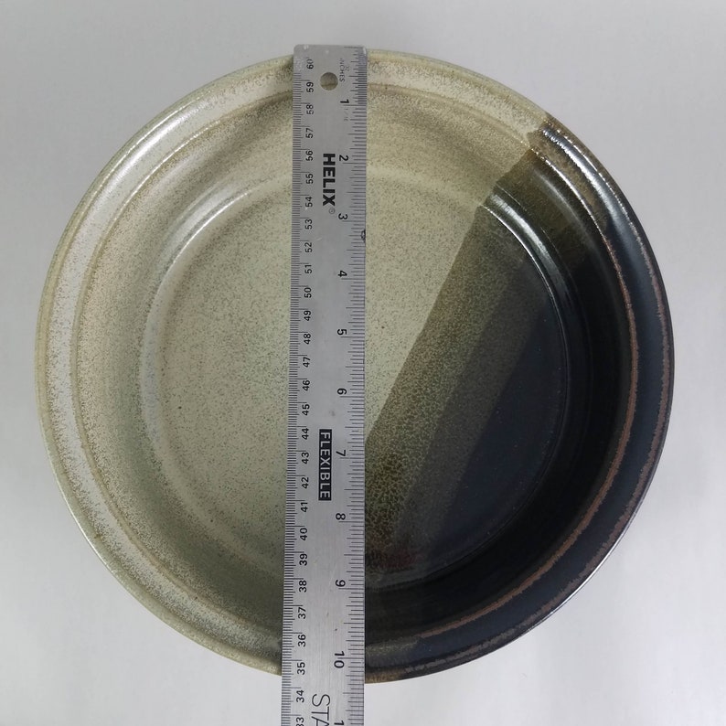 Pu&ograve; includere: Una ciotola rotonda in ceramica con un design bicolore. Il lato sinistro &egrave; beige chiaro, e il lato destro passa dal verde oliva al marrone scuro. Un righello mostra che la ciotola ha un diametro di circa 25 cm.