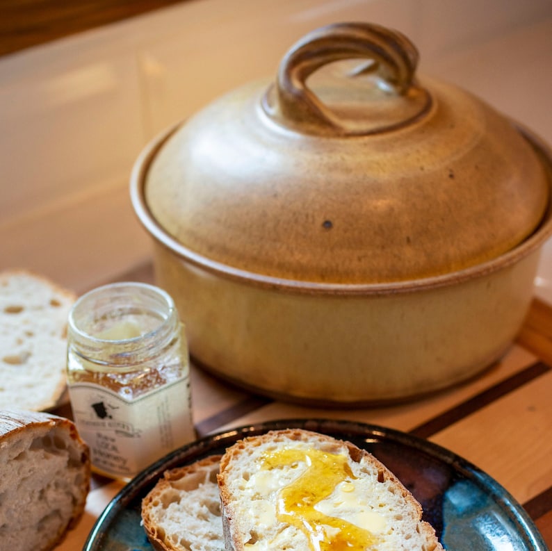 May include: A close-up of a ceramic bread cloche with a lid, a jar of honey, and two slices of bread with honey on a blue and brown plate.