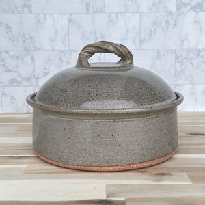 May include: A gray ceramic casserole dish with a lid. The lid has a twisted handle. The dish is round with a slightly textured surface and sits on a reddish-brown base. The background is a light gray and white marble-like pattern.