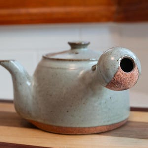 May include: A ceramic teapot with a light green speckled glaze and a side handle. The teapot features a rounded body, a matching lid, and a spout on the opposite side of the handle. The base and the end of the handle show exposed brown clay.