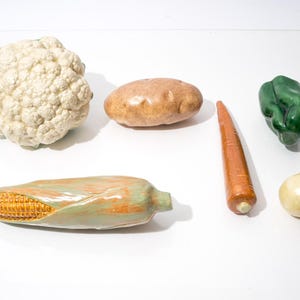 Puede incluir: Un surtido de verduras de cerámica que incluye coliflor, patata, zanahoria, pimiento verde, cebolla y mazorca de maíz. Las verduras están dispuestas sobre una superficie blanca.
