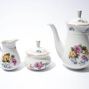 May include: A white porcelain tea set featuring a teapot with lid, a creamer, and a sugar bowl. Each piece is decorated with colorful floral designs in pink, yellow, and blue, with gold trim.