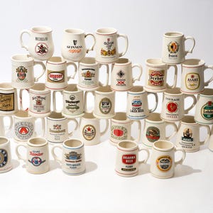 May include: A large collection of cream-colored ceramic beer mugs stacked in a pyramid formation. Each mug features a unique colorful logo and brand name from various international breweries, including Guinness, Asahi, Foster's, and Harp.