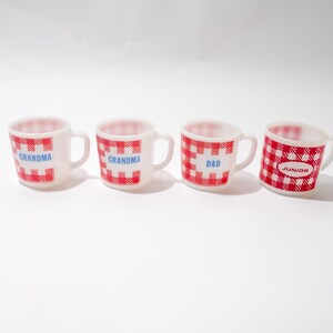 May include: Four white ceramic mugs with red and white checkered patterns and blue text. The mugs read "Grandma", "Grandma", "Dad", and "Junior".