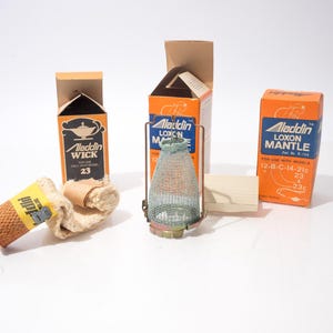 May include: An assortment of Aladdin brand lamp parts, including wicks, mantles, and a metal and mesh lantern. The boxes are orange and brown with the Aladdin logo and product information. The wick is labeled "23".