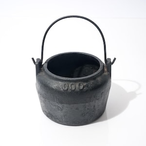 May include: A small, black cast iron pot with a handle. The pot has a rounded body and a wide opening. The handle is made of metal and is attached to the pot with two small brackets. The pot has the number "000" embossed on the side.