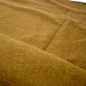 Puede incluir: Primer plano de una tela doblada en un cálido color marrón dorado. El material tiene un aspecto suave y texturizado con sutiles variaciones tonales, lo que sugiere una textura afelpada o lanosa. Los pliegues crean sombras.