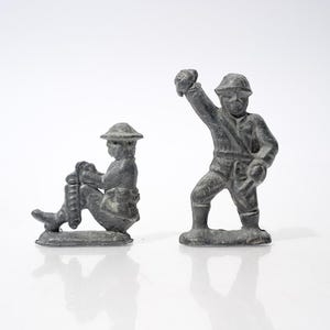 May include: Two gray metal toy soldiers. One is seated, holding a weapon. The other is standing with an arm raised. Both figures wear helmets and uniforms, reminiscent of historical military attire.