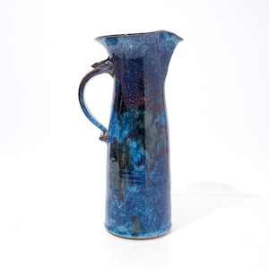 May include: A tall ceramic pitcher featuring a glossy, mottled glaze in shades of deep navy, royal blue, and hints of dark violet. The vessel has a curved handle on the side, a flared rim, and a distinct pouring spout at the top edge.