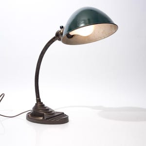May include: An antique desk lamp with a dark green, dome-shaped shade and a flexible, ribbed neck. The base is a dark bronze color. The lamp is turned on, illuminating the interior of the shade. A power cord extends from the base.