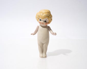 Könnte beinhalten: Eine Vintage-Porzellanpuppe mit blonden, lockigen Haaren. Die Puppe hat einen hellen Teint, rosige Wangen und zarte Gesichtszüge. Ihre Arme und Beine sind beweglich, und sie steht auf einer weißen Oberfläche.